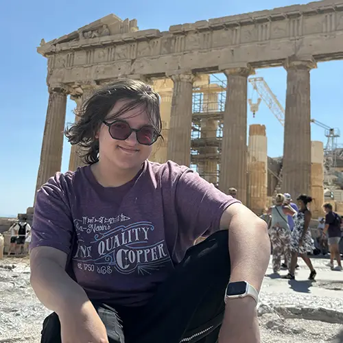 A student in a purple shirt and wearing sunglasses crouching for a photo in front of the Parthenon