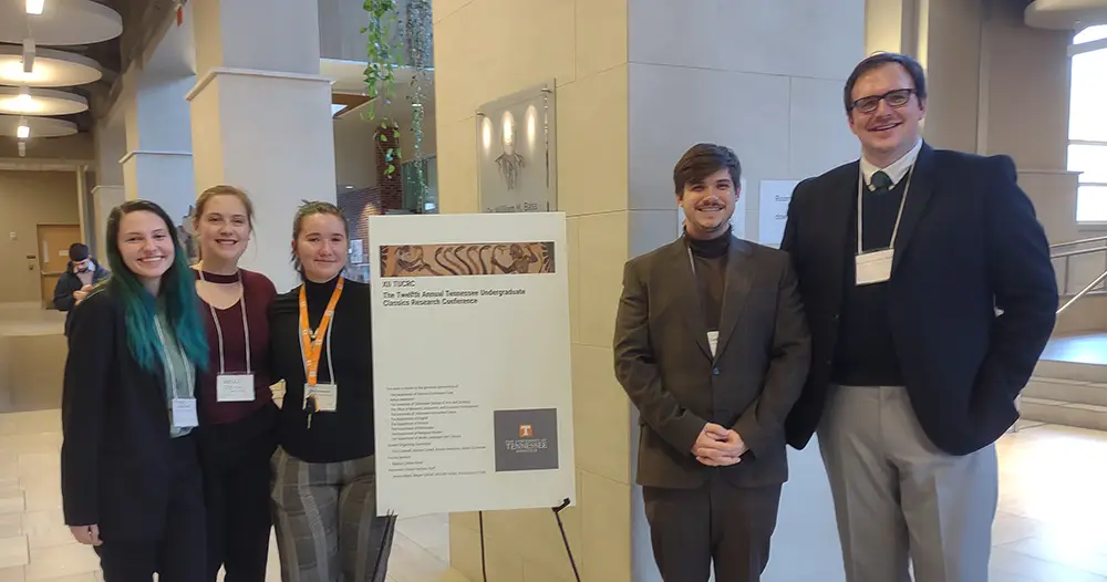 A group photo of five students around a sign and with lanyards and nametags around their necks