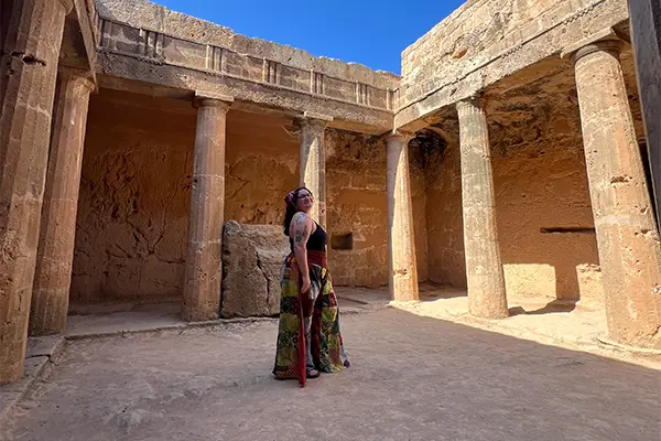 Gabby standing in an ancient stone courtyard with tall Doric columns and weathered walls, likely part of the Tombs of the Kings archaeological site in Paphos, Cyprus.